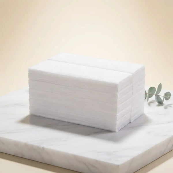 Paire de bandes de cire épilatoire non tissé blanches empilées sur une plaque marble, décor minimal avec brin de feuilles vertes.
